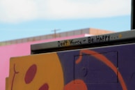 A street art piece featuring a vibrant and abstract design in purple and orange with an encouraging message written on a metal bar. The message reads 'Don't Worry - Be Happy...' accompanied by a small smiley face. In the background, a pink wall and a hint of blue sky are visible.