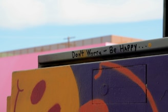 A street art piece featuring a vibrant and abstract design in purple and orange with an encouraging message written on a metal bar. The message reads 'Don't Worry - Be Happy...' accompanied by a small smiley face. In the background, a pink wall and a hint of blue sky are visible.