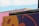 A street art piece featuring a vibrant and abstract design in purple and orange with an encouraging message written on a metal bar. The message reads 'Don't Worry - Be Happy...' accompanied by a small smiley face. In the background, a pink wall and a hint of blue sky are visible.
