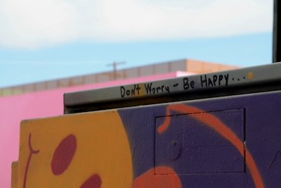 A street art piece featuring a vibrant and abstract design in purple and orange with an encouraging message written on a metal bar. The message reads 'Don't Worry - Be Happy...' accompanied by a small smiley face. In the background, a pink wall and a hint of blue sky are visible.