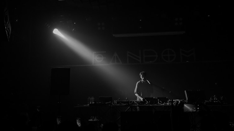 A person stands behind a DJ booth in a dark setting with a spotlight highlighting them. Equipment like mixers and turntables are visible. The word 'FANDOM' is partially visible on the wall in the background.