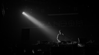 A person stands behind a DJ booth in a dark setting with a spotlight highlighting them. Equipment like mixers and turntables are visible. The word 'FANDOM' is partially visible on the wall in the background.