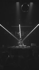 A DJ stands behind a mixer on stage, illuminated by beams of light in a dark club-like setting. A disco ball hangs above, casting subtle shadows. There's a crowd in the foreground, partially visible.