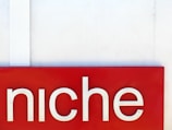 A red rectangular sign with the word 'niche' written in large white letters. The background is a plain off-white wall with a vertical white strip on the left side.