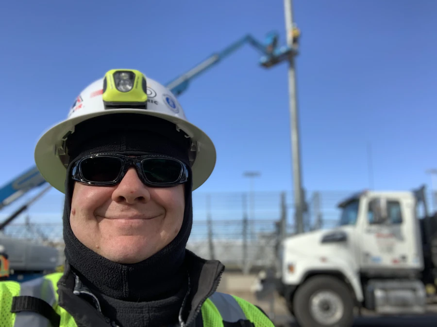 A smiling worker in a white hard hat, headlamp and sunglasses
