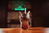 A decadent milkshake is served in a mason jar, topped with whipped cream, chocolate crumbles, and cookies. The jar sits on a wooden table with a blurred background, suggesting a cozy, dimly-lit cafe or restaurant.