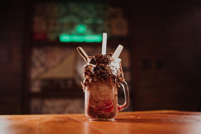 A decadent milkshake is served in a mason jar, topped with whipped cream, chocolate crumbles, and cookies. The jar sits on a wooden table with a blurred background, suggesting a cozy, dimly-lit cafe or restaurant.