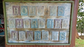 A rustic wooden sign with a weathered appearance, painted with the phrase 'Home is where the heart is'. The sign features a mix of faded blue and brown tones, framed by a green border. The letters are individually painted on square panels, giving it a vintage and handmade feel. Surrounding the sign are glimpses of a chair and an outdoor setting with trees and grass.