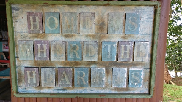 A rustic wooden sign with a weathered appearance, painted with the phrase 'Home is where the heart is'. The sign features a mix of faded blue and brown tones, framed by a green border. The letters are individually painted on square panels, giving it a vintage and handmade feel. Surrounding the sign are glimpses of a chair and an outdoor setting with trees and grass.