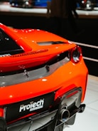 A close-up view of the rear section of a bright orange sports car, featuring a distinctive design with a small horse logo and dual exhaust. The car's sleek body curves and carbon fiber elements are visible, emphasizing its high-performance aesthetics. The environment appears to be a showroom or exhibition setting.