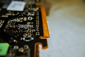 A close-up view of a printed circuit board with intricate patterns and components. The image focuses on the edge of the board with copper contacts visible, resembling those found on a computer memory module or expansion card. The background is softly blurred, emphasizing the detailed electronic pathways and solder points.