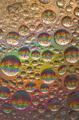 A collection of water droplets resting on a surface, creating a rainbow effect within each droplet. The droplets vary in size and are closely packed together, forming an intricate pattern. The background is a vibrant blend of multiple colors, prominently featuring hues of red, blue, green, and orange, with a subtle reflective quality.