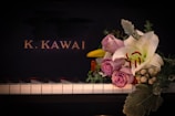 A piano keyboard with a bouquet of flowers placed on it, featuring lavender roses, a white lily, and various greenery. The background is the black surface of the piano with the letters 'K. KAWAI' in gold.