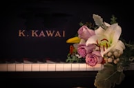 A piano keyboard with a bouquet of flowers placed on it, featuring lavender roses, a white lily, and various greenery. The background is the black surface of the piano with the letters 'K. KAWAI' in gold.