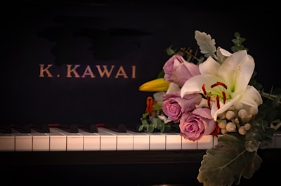 A piano keyboard with a bouquet of flowers placed on it, featuring lavender roses, a white lily, and various greenery. The background is the black surface of the piano with the letters 'K. KAWAI' in gold.
