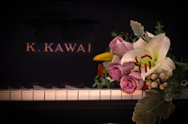 A piano keyboard with a bouquet of flowers placed on it, featuring lavender roses, a white lily, and various greenery. The background is the black surface of the piano with the letters 'K. KAWAI' in gold.
