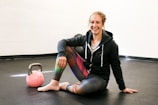 A person is sitting on the floor of a gym or fitness area, smiling. They are wearing a hoodie and colorful leggings, and have one knee up while resting an arm on it. A pink kettlebell is placed on the floor nearby. The setting is simple with a plain wall and dark flooring.