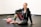 A person is sitting on the floor of a gym or fitness area, smiling. They are wearing a hoodie and colorful leggings, and have one knee up while resting an arm on it. A pink kettlebell is placed on the floor nearby. The setting is simple with a plain wall and dark flooring.