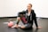 A person is sitting on the floor of a gym or fitness area, smiling. They are wearing a hoodie and colorful leggings, and have one knee up while resting an arm on it. A pink kettlebell is placed on the floor nearby. The setting is simple with a plain wall and dark flooring.