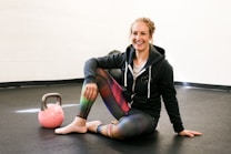 A person is sitting on the floor of a gym or fitness area, smiling. They are wearing a hoodie and colorful leggings, and have one knee up while resting an arm on it. A pink kettlebell is placed on the floor nearby. The setting is simple with a plain wall and dark flooring.
