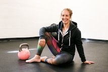 A person is sitting on the floor of a gym or fitness area, smiling. They are wearing a hoodie and colorful leggings, and have one knee up while resting an arm on it. A pink kettlebell is placed on the floor nearby. The setting is simple with a plain wall and dark flooring.