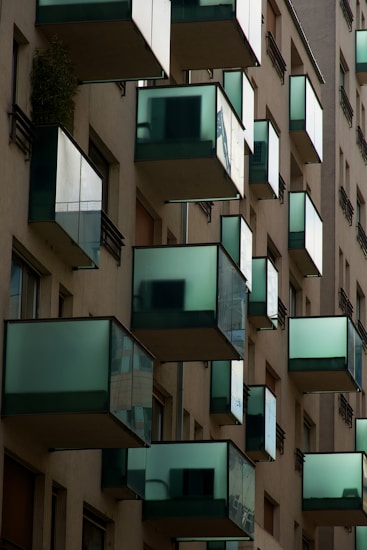 A series of modern balconies jut out from an apartment building. The balconies feature sleek, glass panels that reflect the surrounding environment, giving them a distinctive, contemporary appearance. The building facade is predominantly beige, with clean, straight lines.