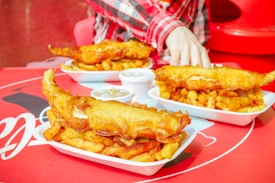 Three trays of golden-brown battered fish and crispy fries are arranged on a red table. A person in a plaid shirt is reaching for some of the food. Small containers of dipping sauce are placed beside the plates.