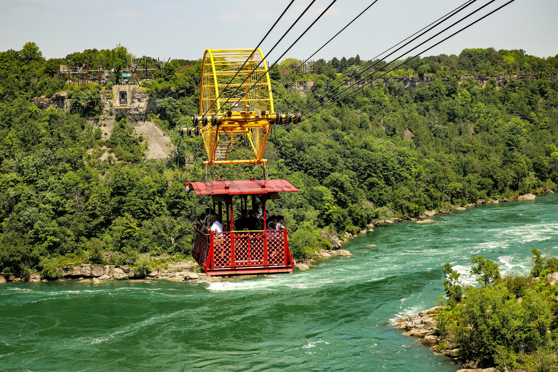 Adventurers crossing a rushing river on a sturdy cable car, with panoramic views of rugged terrain and forests.