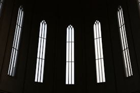 Tall, narrow, gothic-style windows with vertical bars let in streams of light, creating a pattern of shadows and illumination against a dark background.