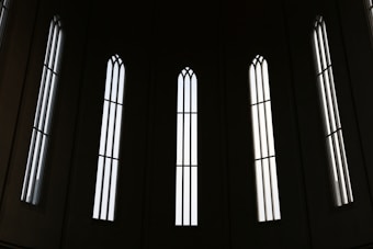 Tall, narrow, gothic-style windows with vertical bars let in streams of light, creating a pattern of shadows and illumination against a dark background.