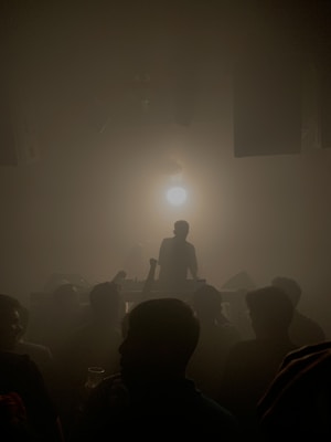 A dark, dimly lit room filled with silhouettes of people facing a DJ booth. A bright light shines behind the DJ, casting shadows on the crowd.