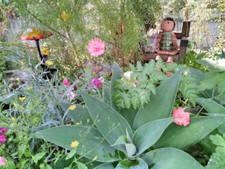 A whimsical garden scene with lush green foliage, pink and yellow flowers, and a colorful mushroom-shaped garden ornament. In the background, there are two whimsical tin can sculptures seated on a small wooden bench, surrounded by a variety of plants.