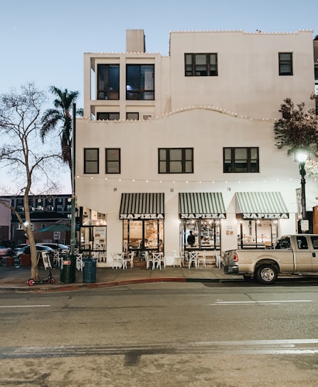 A street view of a building with a white facade, featuring multiple square windows. The ground floor houses a restaurant named 'NAPIZZA', with striped awnings above the glass doors and windows. Small outdoor tables and chairs are set up along the sidewalk. Strings of lights are draped along the building's exterior. A parked pickup truck and a scooter are visible on the street, alongside a lamppost and bare trees under a dusky sky.