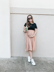 A woman is standing against a light-colored wall, holding a green smoothie in one hand and gently cradling her belly with the other. She is wearing a black top, a peach pleated skirt, white sneakers, and sunglasses. Her expression is relaxed and she is smiling.