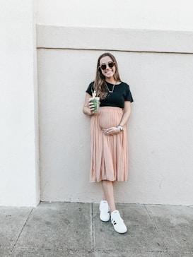 A woman is standing against a light-colored wall, holding a green smoothie in one hand and gently cradling her belly with the other. She is wearing a black top, a peach pleated skirt, white sneakers, and sunglasses. Her expression is relaxed and she is smiling.
