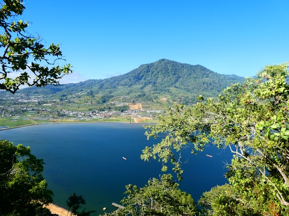A serene landscape with a prominently large lake surrounded by lush greenery. In the background, a sprawling village is nestled at the base of a densely forested mountain. Branches with green leaves frame the image, adding depth and a natural border to the scene.