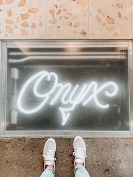 White neon script text spelling 'Onyx' is illuminated on a glass panel. The bottom half shows a person's feet wearing white sneakers, standing on a textured floor. The upper background consists of beige and brown stone tiles.