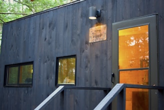 A modern, minimalist building facade made of dark wood with rectangular windows and a warmly lit entrance. A metal plate with the name 'Robert' and a light fixture are mounted near the door.