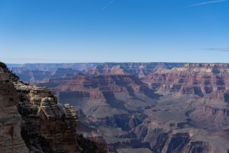 A vast and expansive view of the Grand Canyon showcasing its rugged rock formations and layered cliffs under a clear blue sky. The terrain displays a range of earthy tones with steep drops and jagged edges extending into the distance.