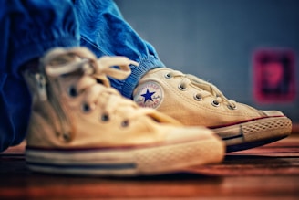 A close-up view of beige high-top sneakers with a distinctive circular logo on the side. The shoes are slightly worn, resting on a wooden surface, and are paired with blue pants.