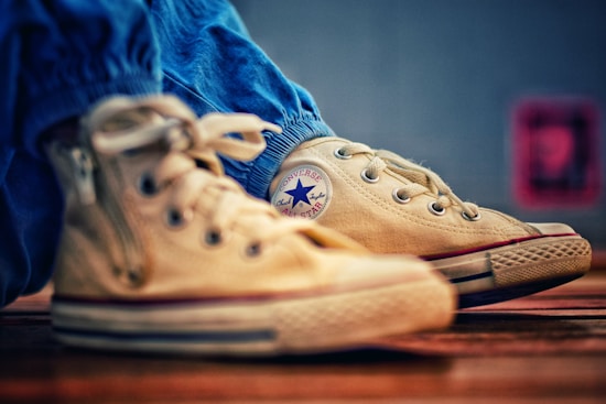 A close-up view of beige high-top sneakers with a distinctive circular logo on the side. The shoes are slightly worn, resting on a wooden surface, and are paired with blue pants.