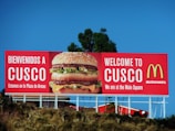 A large McDonald's advertisement billboard stands against a clear blue sky. It features a prominent image of a Big Mac burger, with the text 'BIENVENIDOS A CUSCO' and 'WELCOME TO CUSCO'. The billboard is located near some greenery and appears to be positioned on a raised platform or hill.