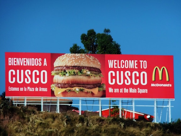A large McDonald's advertisement billboard stands against a clear blue sky. It features a prominent image of a Big Mac burger, with the text 'BIENVENIDOS A CUSCO' and 'WELCOME TO CUSCO'. The billboard is located near some greenery and appears to be positioned on a raised platform or hill.