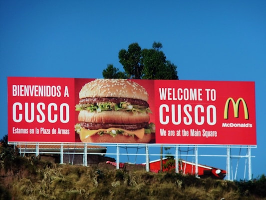 A large McDonald's advertisement billboard stands against a clear blue sky. It features a prominent image of a Big Mac burger, with the text 'BIENVENIDOS A CUSCO' and 'WELCOME TO CUSCO'. The billboard is located near some greenery and appears to be positioned on a raised platform or hill.