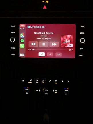 A car infotainment system displaying music information. The screen shows a currently playing song titled 'Sweet but Psycho' by Ava Max, with playback controls visible below the track information. The interface includes various app icons on the left, and temperature and audio controls below.