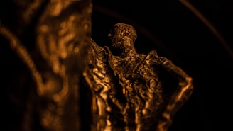 A detailed sculpture crafted in a bronze or metallic color, featuring intricate textures and shadows. The lighting casts deep contrasts, highlighting the sculpted form and its rugged surface.
