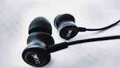 A pair of black earphones with rubber ear tips is placed on a white surface. The earphones have the brand name 'boAt' printed on their sides. The wires are visible and extend from the earphones.