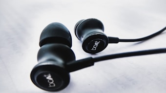 A pair of black earphones with rubber ear tips is placed on a white surface. The earphones have the brand name 'boAt' printed on their sides. The wires are visible and extend from the earphones.