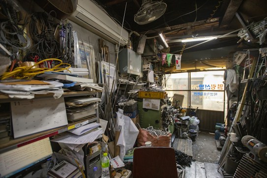 The image depicts a cluttered and cramped workshop or storage area with numerous tools, wires, papers, and equipment scattered around. Shelves are filled with piles of documents and various objects, while the floor is littered with bags, containers, and machinery. Light peeks in through windows partially covered by posters and signs.