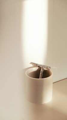A metallic razor placed inside a white ceramic cup, illuminated by soft natural light from the side.